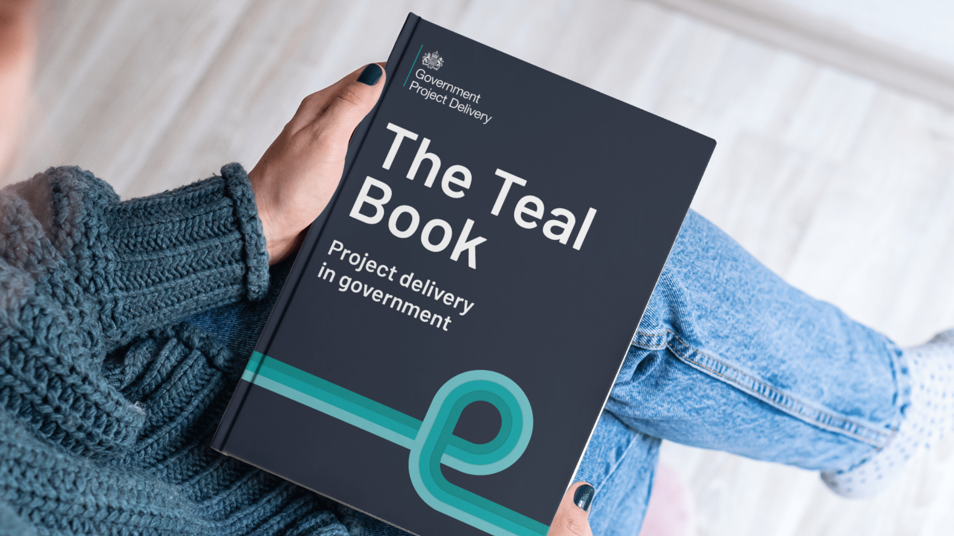 A person holding a book titled “The Teal Book: Project delivery in government,” published by Government Project Delivery. The book has a dark cover with a teal graphic line looping across the bottom.